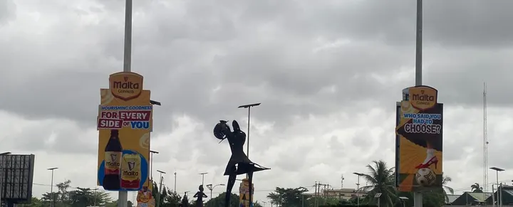 Two Malta Guinness pole die cuts either side of a statue on the Lagos-Ibadan Expressway, Ikeja